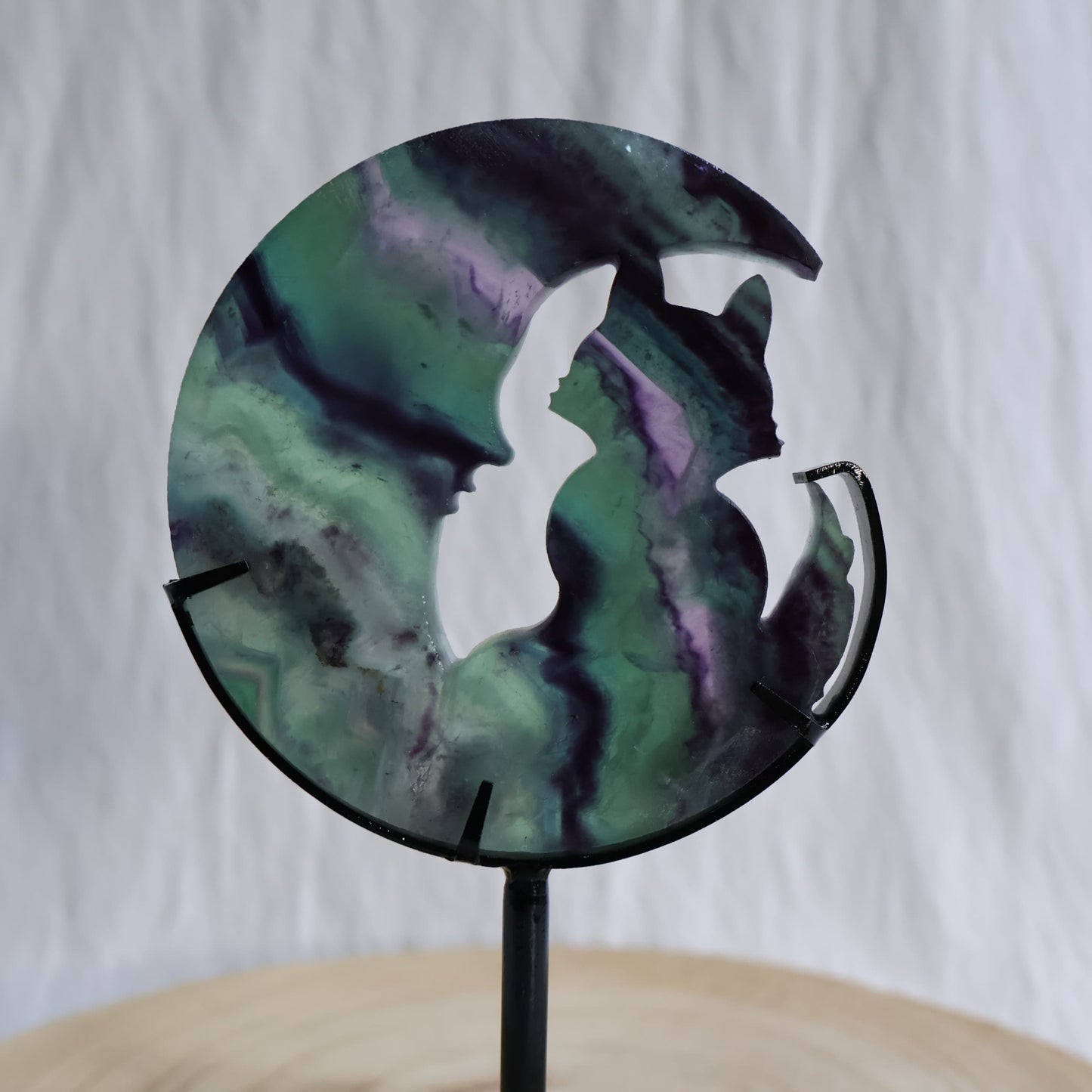 Green and purple Fluorite cat sitting within a crescent moon carving on black metal stand with natural banded colour zoning
