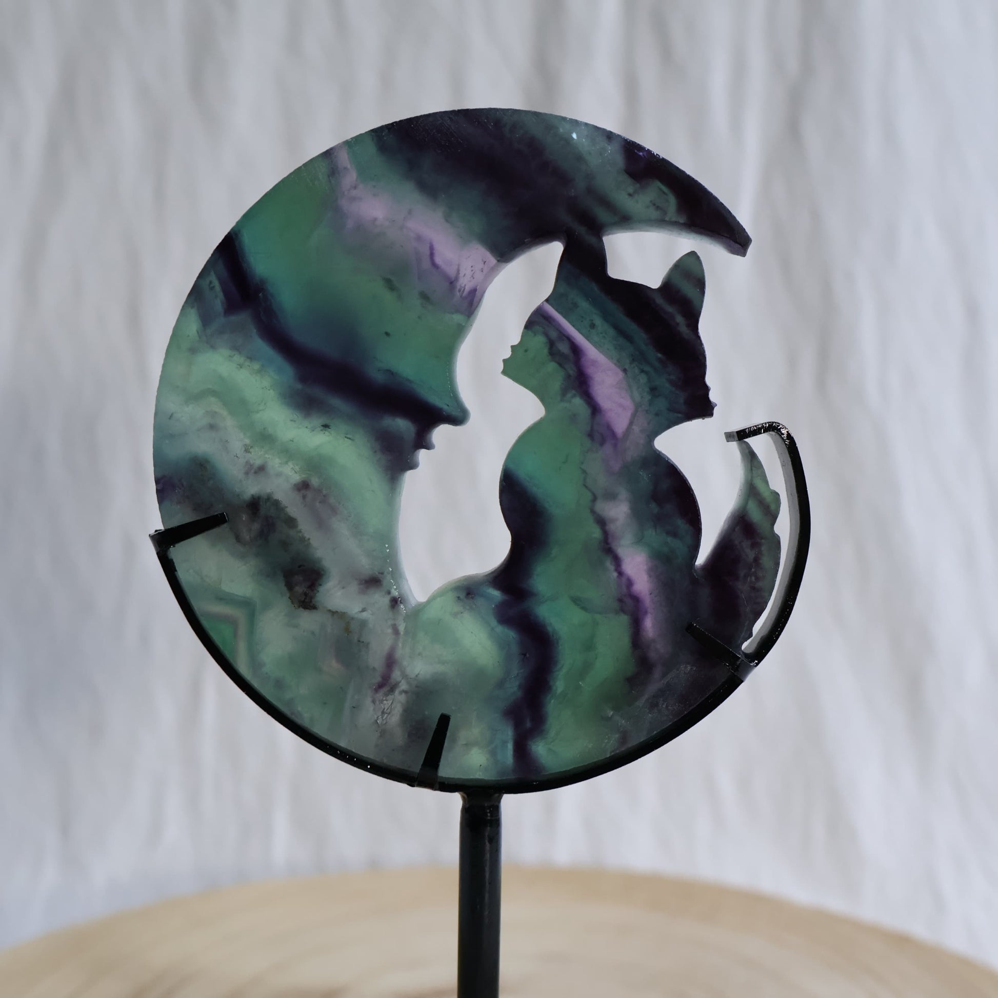 Green and purple Fluorite cat sitting within a crescent moon carving on black metal stand with natural banded colour zoning
