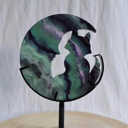 Green and purple Fluorite cat sitting within a crescent moon carving on black metal stand with natural banded colour zoning