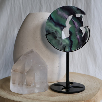 Green and purple Fluorite cat sitting within a crescent moon carving on black metal stand with natural banded colour zoning