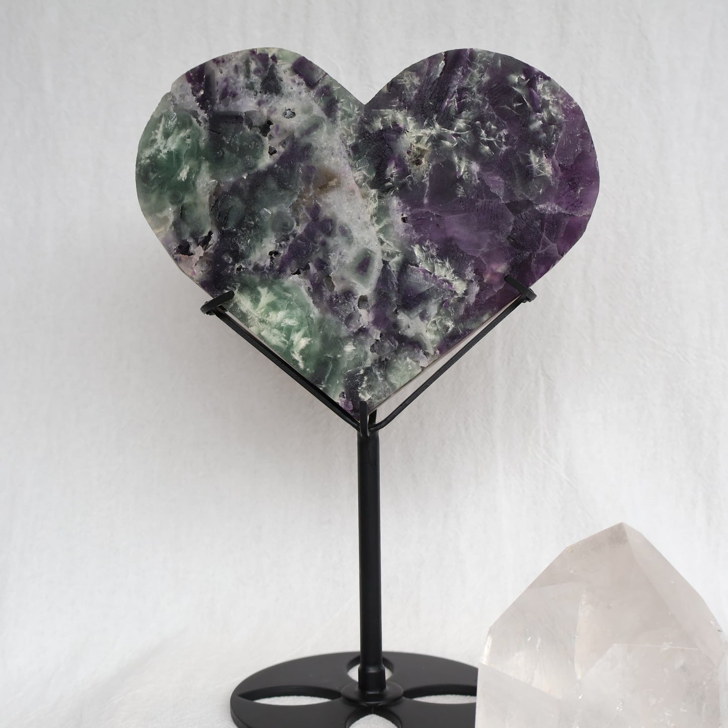 Large purple and green Fluorite heart on black stand with a roughly polished face and contrasting raw crystal reverse, 23cm tall on stand.