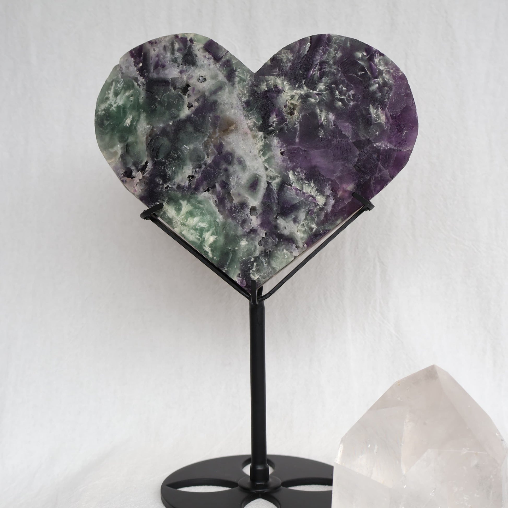Large purple and green Fluorite heart on black stand with a roughly polished face and contrasting raw crystal reverse, 23cm tall on stand.