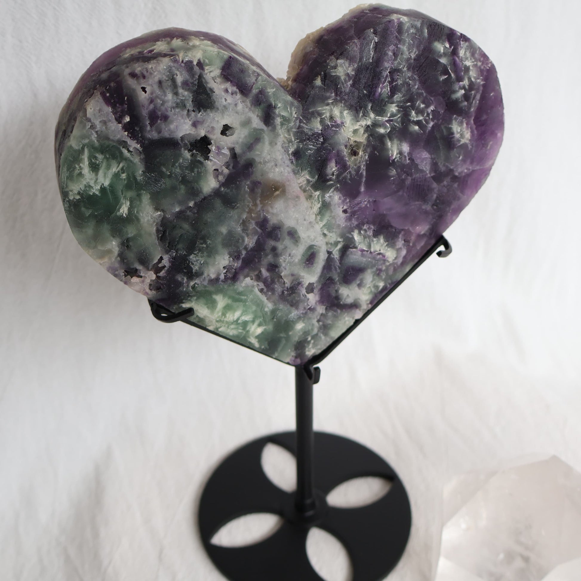 Large purple and green Fluorite heart on black stand with a roughly polished face and contrasting raw crystal reverse, 23cm tall on stand.