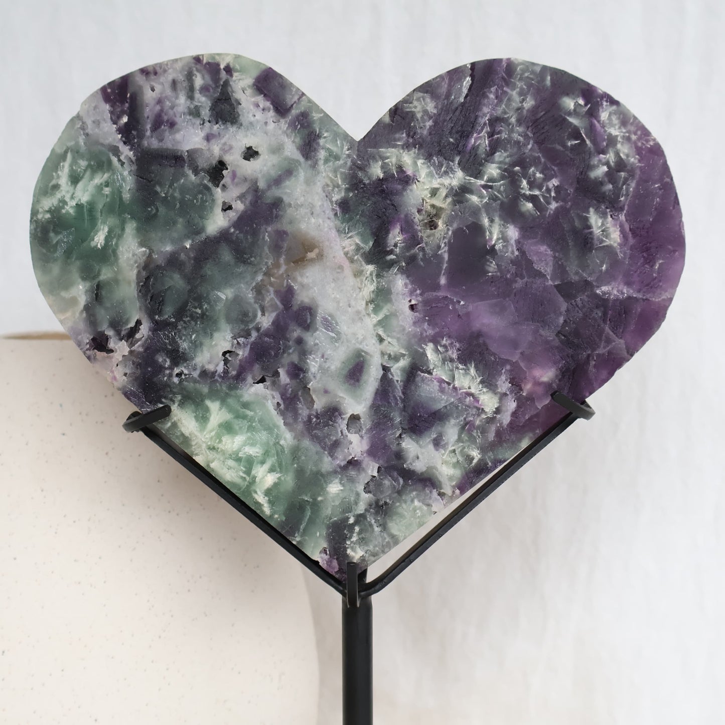 Large purple and green Fluorite heart on black stand with a roughly polished face and contrasting raw crystal reverse, 23cm tall on stand.
