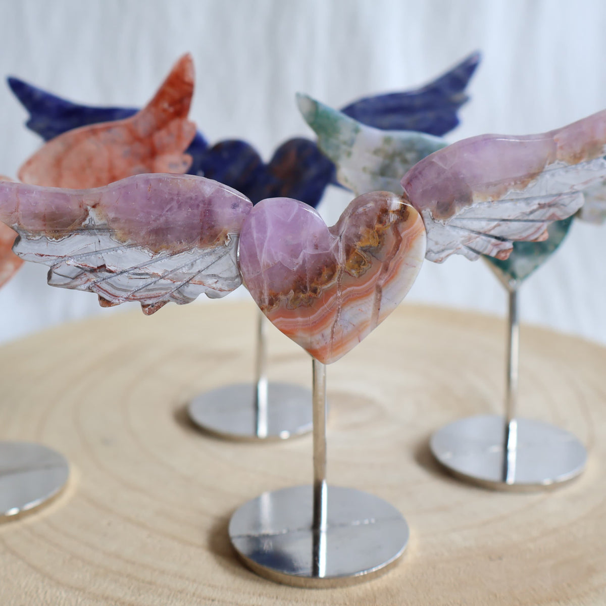 Crystal heart with carved wings in natural Moss Agate, Sodalite, Fire Quartz or Amethyst Agate, displayed upright on a silver-toned stand.